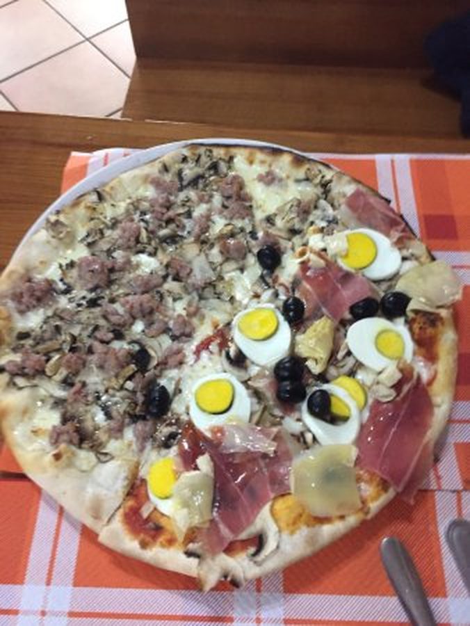 Pizza tonda con prosciutto crudo, uova sode a fette, olive nere e funghi, fotografata dall'alto sul tavolo.
