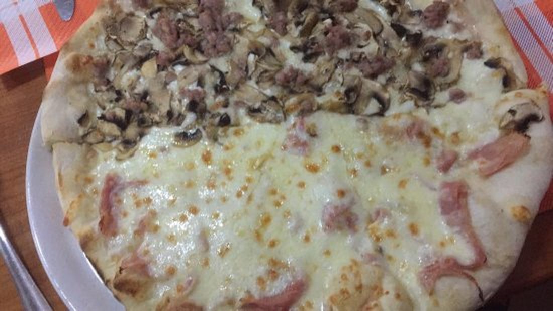 Pizza tonda divisa a metà: da un lato funghi e salsiccia, dall'altro prosciutto cotto e mozzarella.