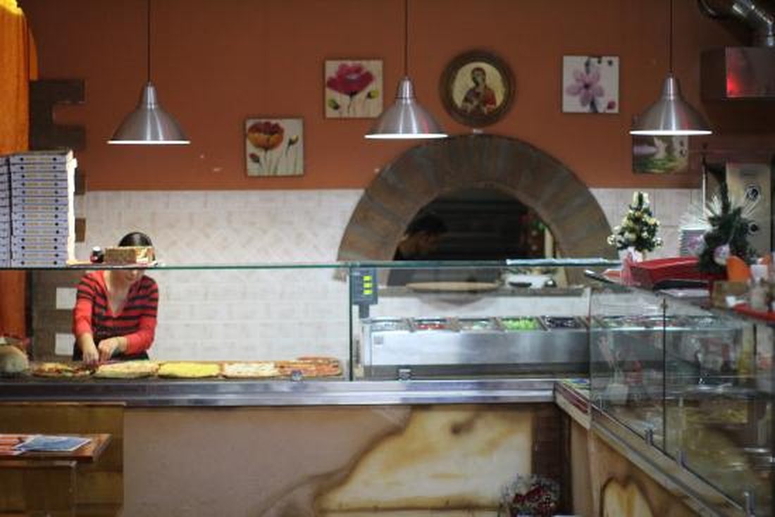Il banco della pizza al taglio di Pizzeria Melody: le teglie in vetrina, il forno a bocca d'arco sul fondo e le scatole per l'asporto impilate a sinistra.